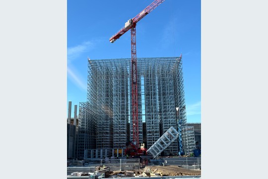 Die Bauarbeiten am Silo-Gebäude schreiten zügig voran: Ein großer Kran hebt gerade einen der vormontierten Regalabschnitte auf seine Einbauhöhe von 45 Metern. (Bildquelle: Dematic)